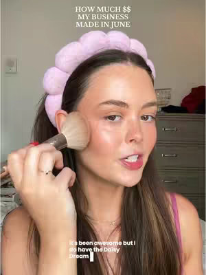 A young woman is applying makeup, holding a makeup brush near her cheek. She's wearing a pink top and a pink headband. Text overlays read "HOW MUCH $$ MY BUSINESS MADE IN JUNE" and a sentence about the Daisy Dream product.