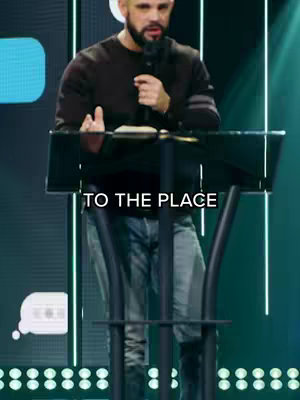 A man with short hair and a beard is standing at a podium holding a microphone. The background is a teal gradient with vertical lights. Text overlays say "TO THE PLACE". The man is wearing a dark long-sleeved shirt and jeans.