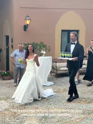 A wedding scene featuring the bride in a white gown, smiling with a drink. A man in a tuxedo carries a tray of drinks in the background, along with other wedding attendees.