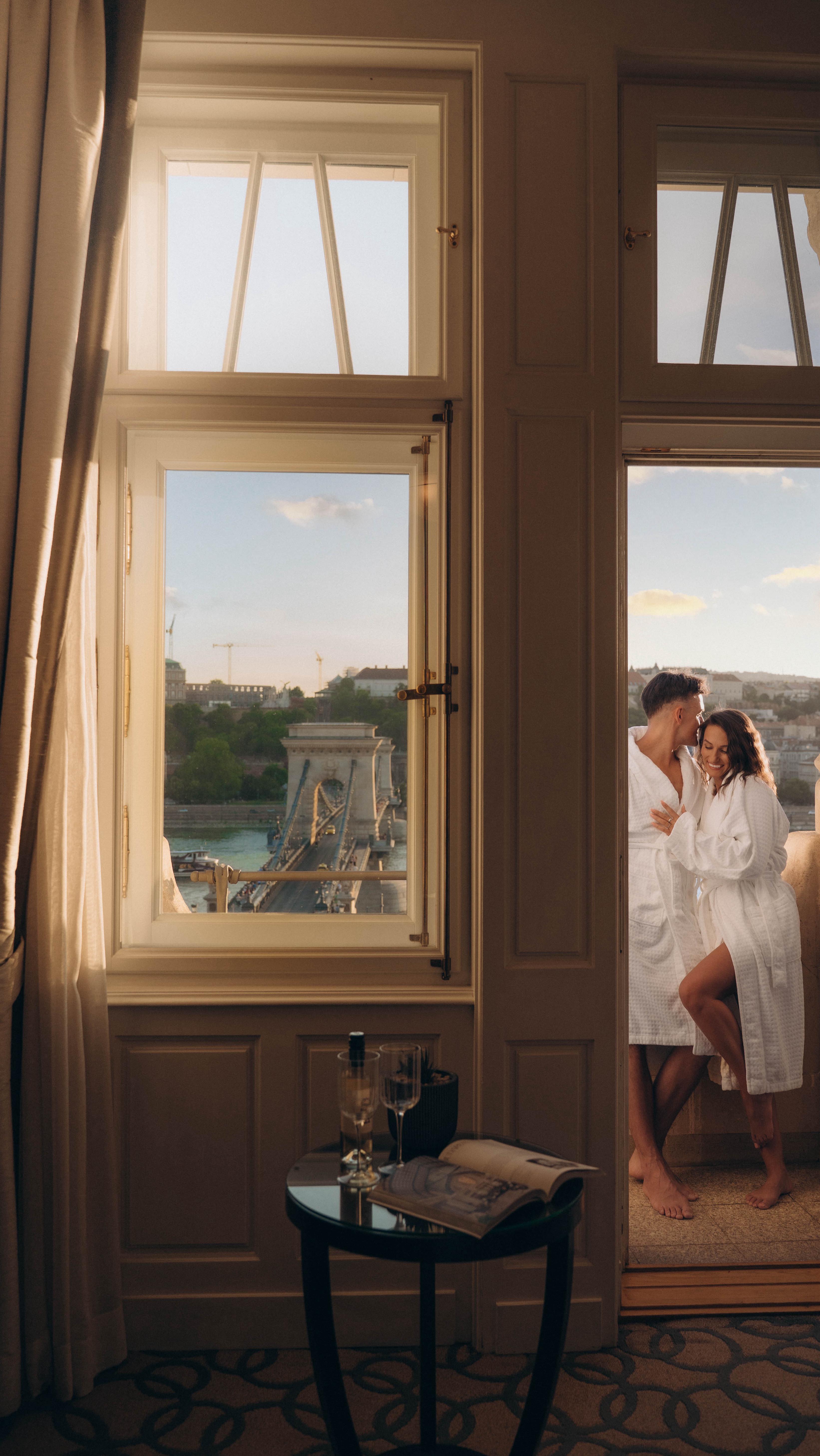 A photograph captures a cozy, well-lit hotel room scene with a focus on a bridge visible through a large window. A couple stands embracing in the doorway to a balcony. The interior features warm tones from the wooden doors and window frames, contrasted by a bright view outside. A round table with a bottle and glasses sits next to the window with an open magazine. The couple is dressed in white robes.