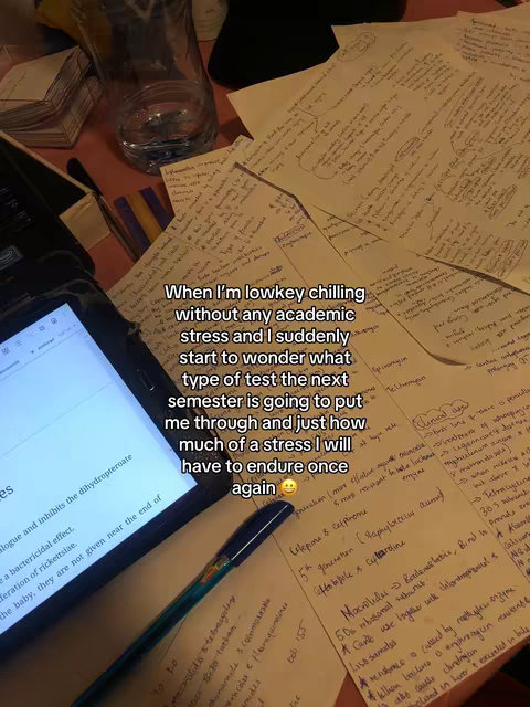 The image showcases a cluttered desk with handwritten notes and a tablet displaying text. A water bottle, pen, and various sheets of paper covered in notes create a study environment. The overall scene suggests an academic, potentially stressful, context.