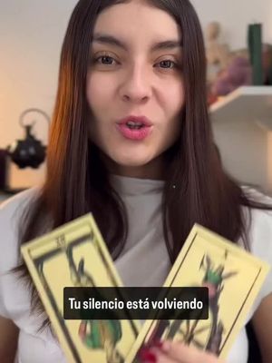 A woman with long brown hair is centered in the frame, looking directly at the viewer and speaking. She is holding two tarot cards, and a transparent overlay at the bottom displays the text: "Tu silencio está volviendo". The cards are a gold color on a black background. The lighting is bright with a slightly warm tone.