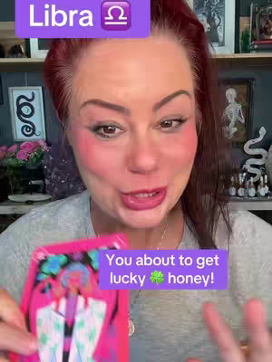 A woman with red hair and makeup is holding up a tarot card. Behind her are shelves and objects. Overlaid onto the image are two text boxes, one with the zodiac symbol for Libra. The other saying, "You about to get lucky honey!"