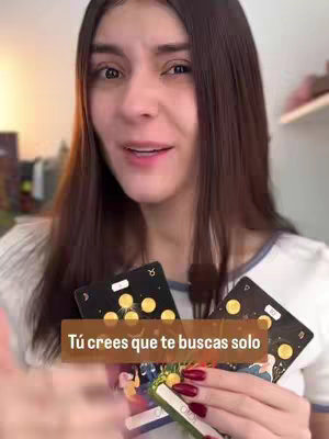 A selfie-style shot features a woman with long, dark hair, holding two tarot cards. She appears to be in the middle of speaking, with a neutral expression. The cards have gold symbols on a dark background. Text overlaid on the image reads, "Tú crees que te buscas solo."