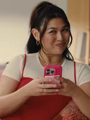 A young woman with dark hair pulled back in a high ponytail, is holding a pink phone in front of her, with a slight smile, wearing a white t-shirt and a red top, gold necklaces and earrings.
