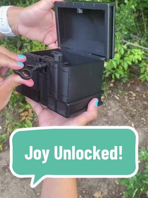 A person holds a black treasure chest, the lid partially open. The chest has a key in the lock. The background shows green foliage and sunlight. Text overlay: "Joy Unlocked!" in a speech bubble.