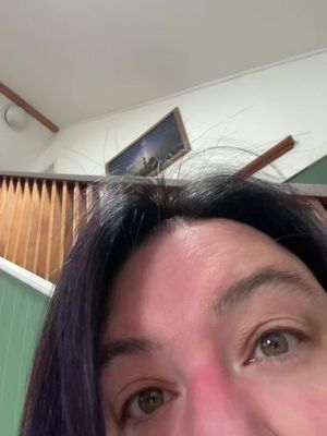 A close-up shot of a person's face, angled slightly downwards. The focus is on the eyes, which are light brown. Above the eyes is dark hair, and the forehead and cheeks appear to be slightly flushed or illuminated. Behind the person, a white ceiling and part of a wooden staircase are visible, with a framed picture.