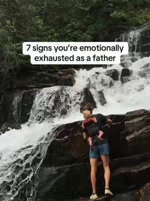 A man wearing a dark shirt, blue shorts, and tan shoes is standing on a rock by a waterfall, with a baby in a carrier on his chest. The waterfall is behind him, and the image is surrounded by lush green trees and vegetation. Text overlayed in a white rectangle says, "7 signs you're emotionally exhausted as a father."