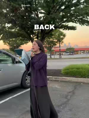 A woman stands in a parking lot holding a microphone, speaking. She is wearing a long black skirt and a dark purple top. The background includes a parking lot, a few cars, trees and a strip mall. The sky is a gradient of orange and blue representing either a sunset/sunrise.