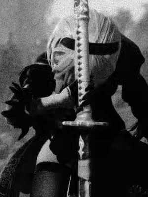 A black and white photograph depicts a character from the game Nier: Automata. The character is mostly in shadow but appears to be wearing a white blindfold and a black outfit. She is holding a large sword with a distinctive design in front of her as the central focus. The image is grainy and has a slightly muted tone.