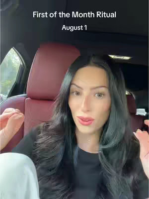 A woman sits in a car, looking at the camera. The video cover has the text "First of the Month Ritual, August 1." The car's interior is visible, with burgundy-colored seats. The woman has long dark hair and is wearing a black top.