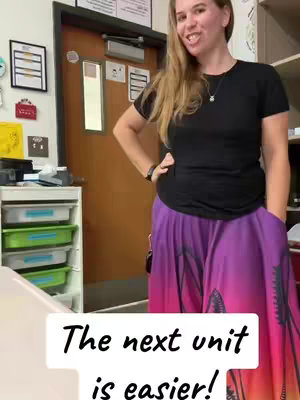 A woman stands in what appears to be a classroom setting, smiling at the camera while leaning against a door. She wears a black t-shirt and a long skirt with vibrant pink and purple colors. The text overlay reads, 'The next unit is easier!'