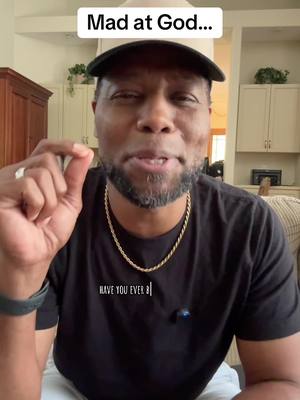 A close-up shot of a Black man wearing a baseball cap and a black t-shirt against a blurred background of a kitchen. He is making a hand gesture and appears to be speaking.