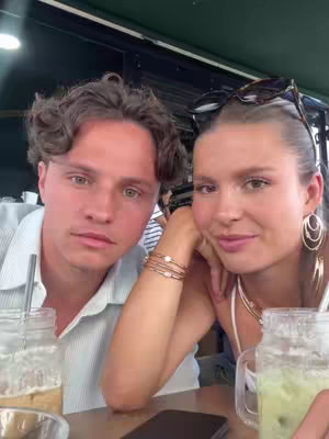 A selfie-style shot of a young man and woman seated at a restaurant table. The man, on the left, has short brown hair and a light blue button-down shirt. The woman, on the right, has long brown hair, sunglasses on her head, and is wearing a white top. They are both smiling and facing the camera. On the table are two drinks in glass jars. The background suggests an outdoor cafe setting.