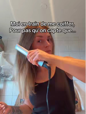 A woman is filming herself in what appears to be a bathroom, holding a hair straightener. Text overlays the image, partially obscured by the woman and her hair. She wears a black tank top and has tattoos on her arm. The background is generic and the lighting appears to be from overhead.