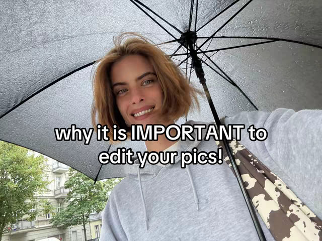A selfie taken under an umbrella on a rainy day. The person holding the umbrella, a woman with short, light-brown hair and a grey hoodie, smiles at the camera. The background shows a building facade and trees. Overlay text in black letters says WHY IT IS IMPORTANT to edit your pics!
