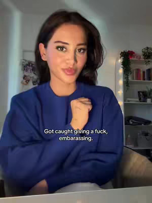 A woman with dark hair and light skin is centered in the frame, wearing a blue sweater. She has a natural, slightly smirking expression, with her left hand raised near her mouth. The background shows a soft-lit room with a white bookshelf and a wall.