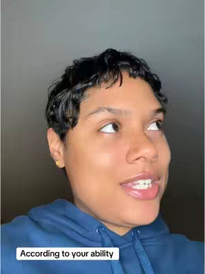A selfie-style shot featuring a person of color with short, dark hair. They are wearing a blue hoodie and looking upward and to the right with a thoughtful, slight smile. The scene is lit with soft lighting and set against a plain, gray background. A text overlay at the bottom reads 'According to your ability'.