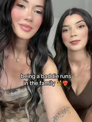 Two young women are positioned closely together, with their faces angled towards the camera. The woman on the left has dark hair with lighter streaks, while the one on the right has dark hair. Both women have fair skin and are wearing tops that show some cleavage. Text overlays are present on the image, with a playful heart emoji.