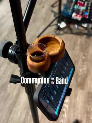 The image displays a close-up of a wooden holder with a small communion cup filled with a dark liquid, mounted on a black stand with a smartphone attached. The smartphone shows a sound mixing interface. The background is blurred, showing stage equipment. The text overlay in white font states 'Communion for the Band'.