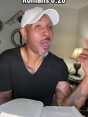 A man with a white cap and a black v-neck shirt is looking at the camera with an open mouth, as if surprised. Behind him is a room with some decorative items and a table lamp. He is holding an open book.