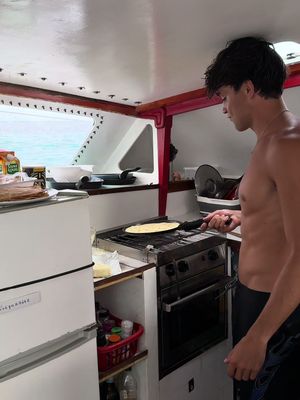 A man in the kitchen area of a boat prepares food on a stove top, wearing no shirt. The kitchen is white with red accents and a window that looks out to the ocean is behind him.