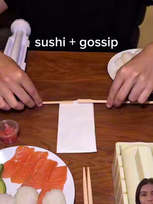 A person is making sushi on a wooden table. The image is a medium shot with the person's hands centered, actively arranging sushi components. The layout includes a white plate on the left with sliced salmon, a plate of white cooked rice, small containers of pickled ginger, chopsticks, a sushi rolling mat, and a white sushi press.
