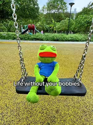 A plush frog sits on a swing in a park, positioned in front of a blurred green background of trees, foliage, and playground equipment. The frog has bright green skin, a blue shirt, and a red mouth. The text 'Life without your bobbies' is written at the bottom in white