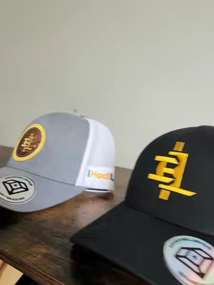 The image showcases two baseball caps on a wooden surface. The cap on the left is grey with a white mesh back and a circular patch featuring a logo. The cap has a sticker on the brim. The cap on the right is black with an embroidered yellow logo and also has a sticker on the brim. The background is a plain off-white wall, and the lighting appears natural.