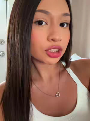 A close-up shot of a young woman with long, dark hair, brown eyes and full lips, appearing to speak directly to the camera. She wears a white top and a gold heart pendant necklace. The lighting is soft and diffused.