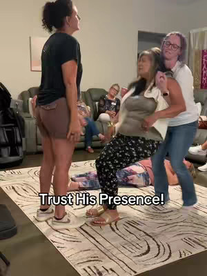 The image captures a group of women in an indoor setting. Two women are supporting another woman who appears to be overcome with emotion, perhaps in a religious context. Other women are in the background, some seated and observing. The scene takes place on a patterned rug, with a couch and other furniture visible. Additional text overlay 'Trust His Presence!' is written in bold white font at the bottom of the image, contrasting against the dark background.