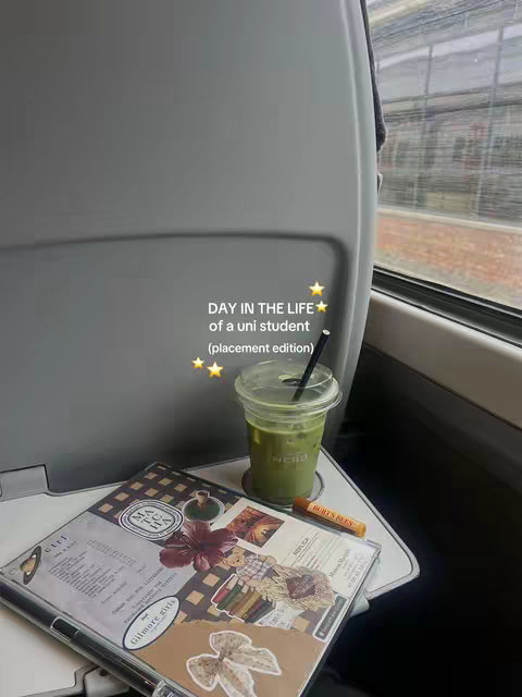 A photo taken inside a train. The main focus is on the items placed on a tray table. A cup of green drink with a black straw, a notebook, and a lip balm are visible. In the background, there is a blurred view through the train window. Text overlay on the image are the words 'DAY IN THE LIFE' above the words 'of a uni student (placement edition)’ and 3 stars.
