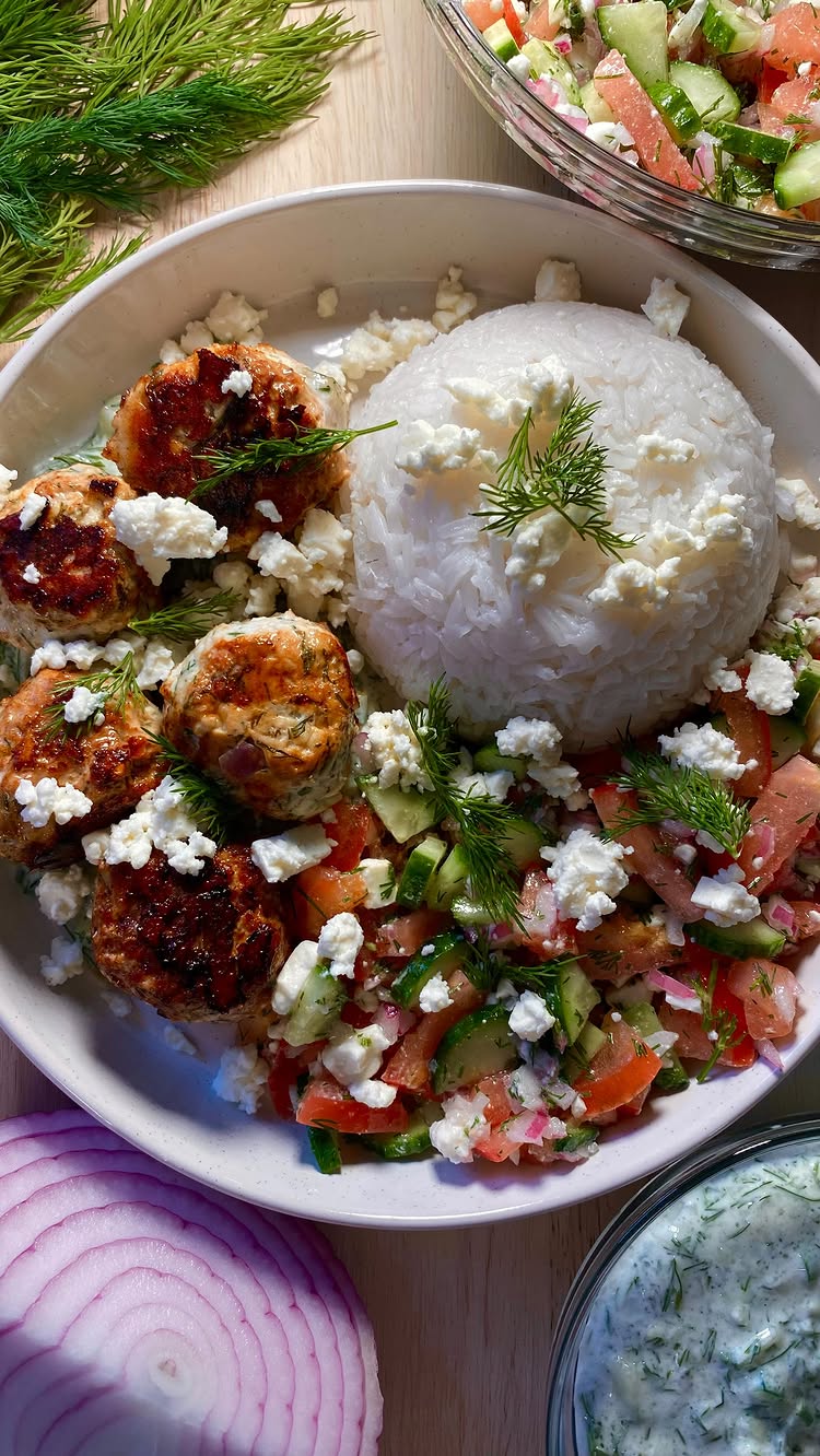 The image showcases a beautifully composed Mediterranean Meatball Bowl.  A large, off-white bowl holds a generous portion of fluffy white rice, topped with crumbled white feta cheese. Around the rice are several golden-brown, round meatballs and a colorful salad of diced tomatoes, cucumbers, red onions, and herbs, also sprinkled with feta.  A small clear bowl with a creamy white dip is also visible.  The food is arranged on a light-colored wooden surface, with a sprig of fresh dill as a garnish.  The overall composition is appealing and food-focused, with a top-down perspective.