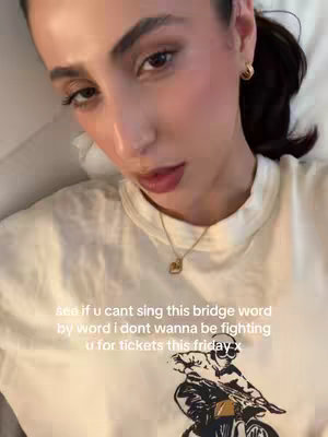 A selfie-style shot of a person lying down on what appears to be a bed, looking at the camera. The person has dark hair pulled back, visible makeup. The lighting is soft. Overlayed text appears over the person, with a graphic on the clothing worn.