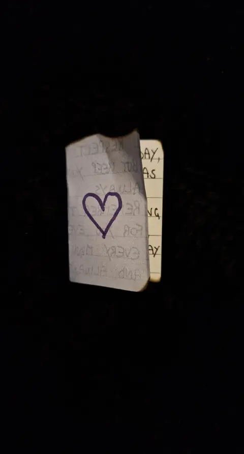 A black background with a torn, crumpled piece of paper in the foreground. A drawn purple heart is visible on the paper. To the right, a yellow sticky note with handwritten text.