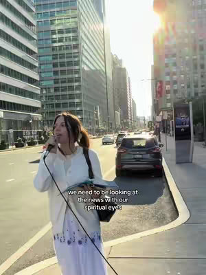 A woman is standing on a sidewalk speaking into a microphone. She is wearing a white cardigan, a white blouse, and a long, light-colored skirt with a blue floral pattern. Behind her is a city street with tall buildings on either side. Sunlight bathes the scene.