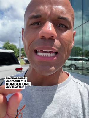 A bald man with a gray t-shirt is speaking toward the camera. He has his mouth open, showing his teeth. The background appears to be near a building with windows and some cars. A text overlay is visible in the lower left corner.