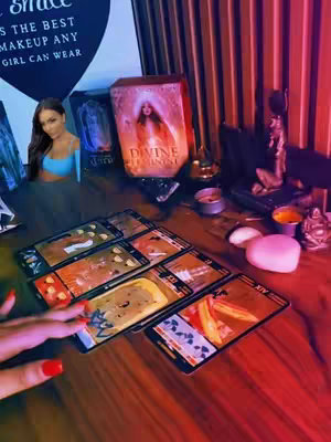The image displays a tarot card reading with a woman's hand selecting cards. The woman is seen in a small inset, in the background are tarot card boxes and candles casting a warm, mystical glow. The lighting is a mix of warm red and cool purples.