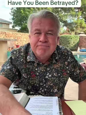 A middle-aged man with gray hair and a floral shirt is shown from the chest up, looking at the camera with a neutral expression. The background includes outdoor elements like trees, foliage, and a glimpse of a swimming pool, suggesting a backyard or patio setting. A partially visible book or notepad is held in front of him. Text overlay at the top says "Have You Been Betrayed?"
