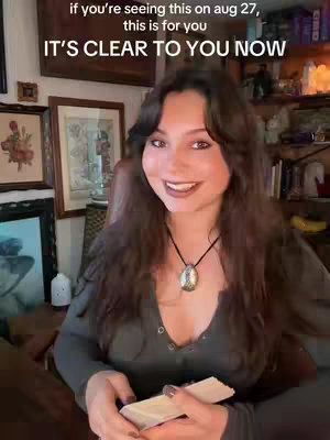A woman with long brown hair is seated, smiling with a tarot reading in front of her, framed against a background of artwork and a bookshelf.
