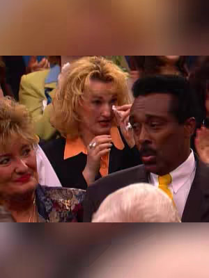 A screenshot from a performance, likely in a church or auditorium. A woman in foreground is dabbing her eye, appearing to be crying. A Black man stands near her, and several other people are visible in the background.