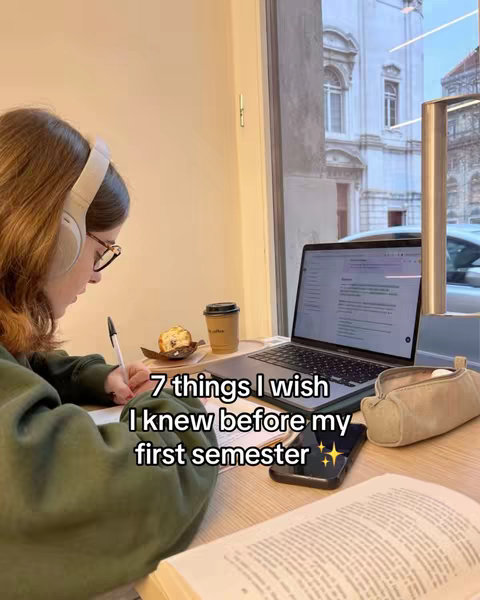 A young woman with long brown hair sits at a wooden table, studying. She wears glasses and headphones, and is writing with a pen on a notepad. A laptop is open in front of her, and a cup of coffee and a pastry are also on the table. The window behind her shows the blurred street scene. Text overlayed is '7 things I wish I knew before my first semester'.