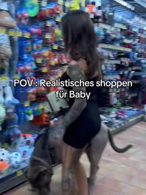 A woman holding a dog on a leash is in a pet store with shelves of products in the background. Text overlay reads: "POV: Realistisches shoppen für Baby".