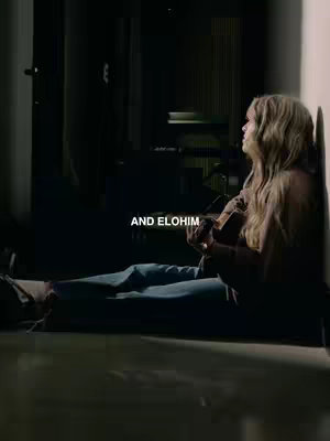A woman is seated on a floor with her back against a wall, in a dimly lit space. She is holding a guitar, and her head is tilted upward, as if looking towards a light source just outside the frame. Text overlayed on the image reads 'AND ELOHIM'. The overall mood is contemplative and serene.