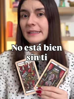 A woman with long dark hair is centered in the frame, slightly angled towards the camera. She wears a patterned blouse and has fair skin with visible makeup. She is holding two tarot cards with colorful illustrations depicting a devil and a person in a jar. The text overlayed says 'No está bien sin ti'. Behind her is a blurred background of shelves and objects.
