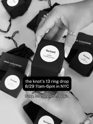 The image is a grayscale overhead shot capturing multiple small black boxes with white circular labels. The labels display the text 'the knot' and 'RING CONCIERGE'. The foreground shows a hand holding a box open, revealing a ring, with another ring visible on the hand. Additional boxes are scattered around the hand. Text overlayed reads: 'the knot's 13 ring drop 8/29 11am-6pm in NYC' and 'baby, let the games begin'.