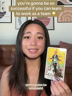 A young woman with long, dark hair, wearing a coral-colored top, smiles into the camera while holding a tarot card. Above her, a text overlay states, “you’re gonna be so successful if you can learn to work as a team” in bold, light-brown lettering with a gold star. Below, the text "you're gonna be successful" in a similar style. The background includes a blurred living room scene, with framed artwork. The tarot card features an image of a person riding a motorcycle, in shades of yellow, blue, and black.
