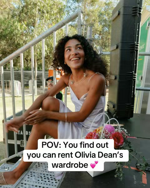 The cover image features a woman with curly dark hair sitting on metal stairs outdoors, smiling and looking upwards. She is wearing a light-colored dress. A flower arrangement in a white basket sits to her right. Text overlay shows 'POV: You find out you can rent Olivia Dean's wardrobe' with two pink hearts.