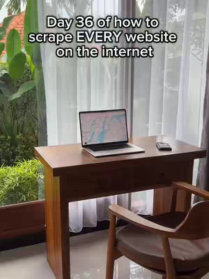 A laptop sits on a wooden desk near a window with sheer curtains. The laptop screen displays a map, and a smartphone rests beside it. The setting suggests a work-from-home environment with natural light and outdoor greenery visible through the window. The text overlay describes the video as being about scraping websites.