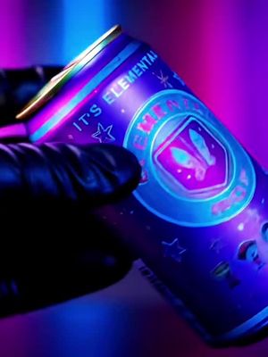A close-up shot of a beverage can held by a gloved hand. The can has a vibrant purple and blue color scheme with glowing accents. Text and a logo are visible on the can. The lighting suggests a stylized environment, possibly with blacklight.